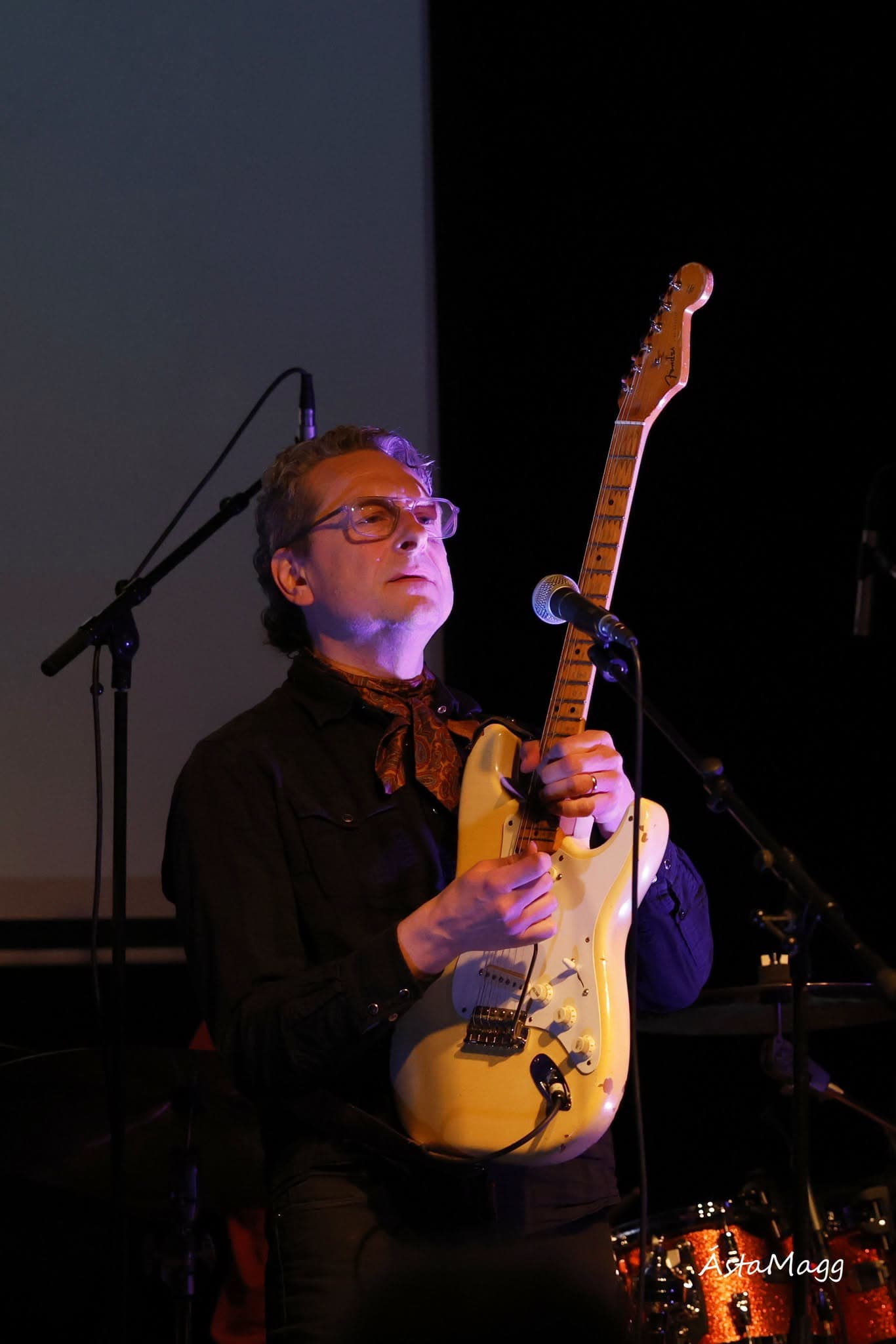 Gummi P plays an electric guitar on a dimly lit stage